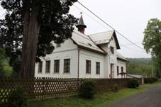 Přízemní běle zbarvená budova se šikmou střechou zakončenou malou zvoničkou. Vlevo před budovou roste listnatý strom